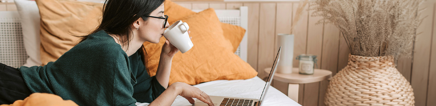 Woman on her bed looking at a laptop while she drinks coffee