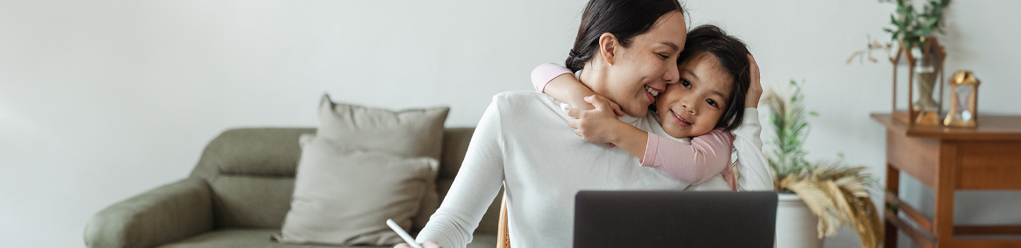 A mother and daughter hugging while the mother sits in front of a laptop